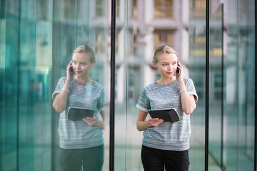 Young beautiful business woman in modern glass interior speaking on the phone and using tablet