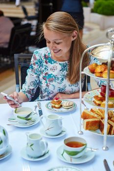 Beautiful young woman enjoying afternoon tea with selection of fancy cakes and sandwiches in a luxury Parisian restaurant. Girl is checking her mobile phone
