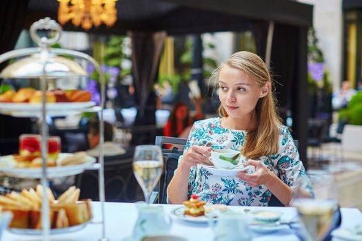 Beautiful young woman enjoying afternoon tea with selection of fancy cakes and sandwiches in a luxury Parisian restaurant