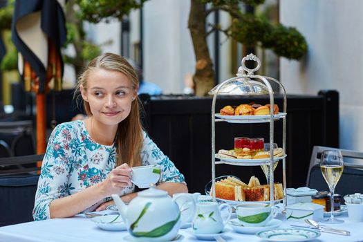 Beautiful young woman enjoying afternoon tea with selection of fancy cakes and sandwiches in a luxury Parisian restaurant