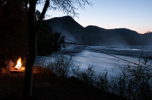 A campfire at sunset next to the Kunene River at the top of the Epupa waterfalls