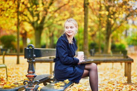 Beautiful young woman in the Luxembourg garden of Paris on a fall day, drinking coffee to go and reading book