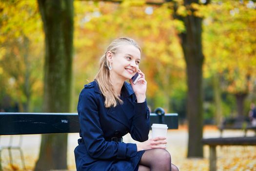 Beautiful young woman in the Luxembourg garden of Paris on a fall day, drinking coffee to go and using mobile phone