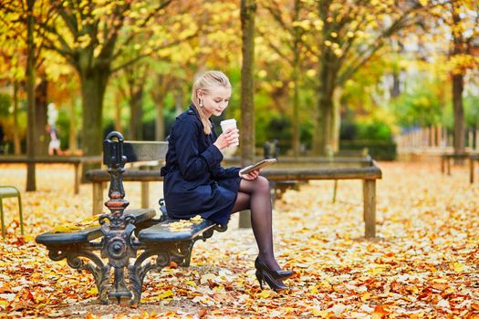 Beautiful young woman in the Luxembourg garden of Paris on a fall day, drinking coffee to go and reading book