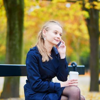 Beautiful young woman drinking coffee and speaking on the phone in the Luxembourg garden of Paris on a fall day