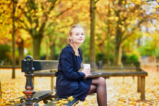 Beautiful young woman in the Luxembourg garden of Paris on a fall day, drinking coffee to go and reading book