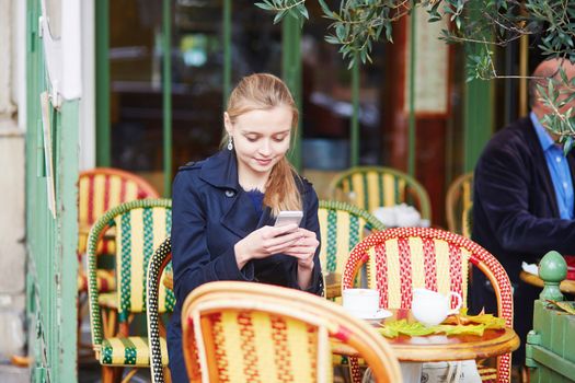 Beautiful young woman drinking hot chocolate in Parisian outdoor cafe and taking photo with her mobile phone