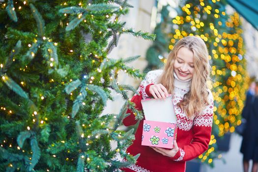 Cheerful young woman on a street of Paris decorated for Christmas, unpacking Christmas present