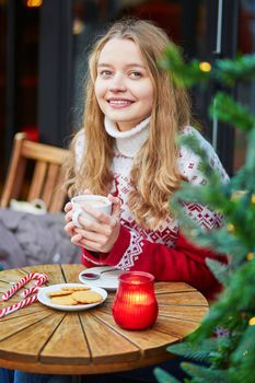 Cheerful young woman drinking hot chocolate in a cozy outdoor Parisian cafe decorated for Christmas
