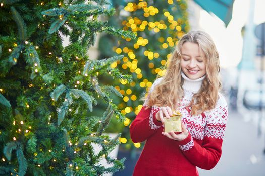 Cheerful young woman on a street of Paris decorated for Christmas, unpacking Christmas present