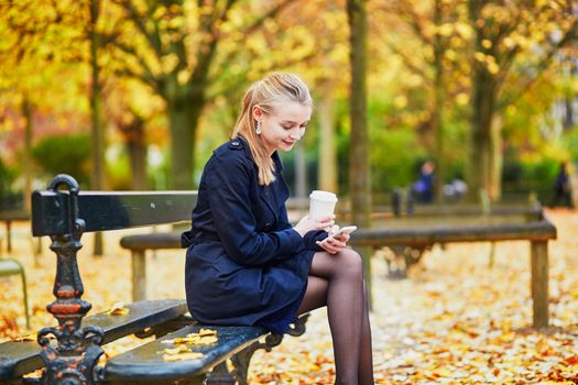 Beautiful young woman in the Luxembourg garden of Paris on a fall day, drinking coffee to go and using mobile phone