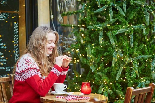 Cheerful young woman drinking hot chocolate in a cozy outdoor Parisian cafe decorated for Christmas
