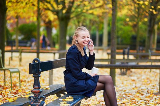 Beautiful young woman in the Luxembourg garden of Paris on a fall day, drinking coffee to go and using mobile phone