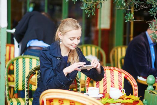 Beautiful young woman drinking hot chocolate in Parisian outdoor cafe and taking photo with her mobile phone