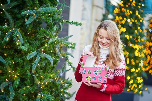 Cheerful young woman on a street of Paris decorated for Christmas, unpacking Christmas present