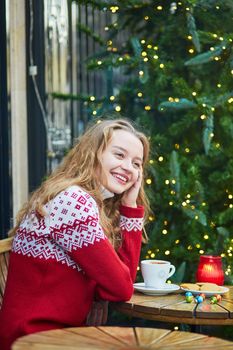 Cheerful young woman drinking hot chocolate in a cozy outdoor Parisian cafe decorated for Christmas