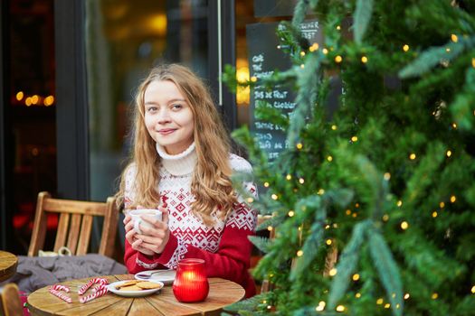 Cheerful young woman drinking hot chocolate in a cozy outdoor Parisian cafe decorated for Christmas