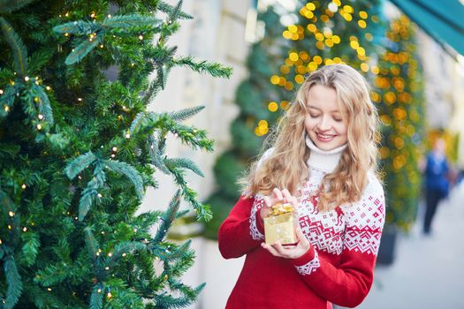 Cheerful young woman on a street of Paris decorated for Christmas, unpacking Christmas present