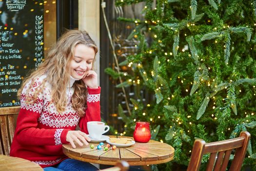 Cheerful young woman drinking hot chocolate in a cozy outdoor Parisian cafe decorated for Christmas