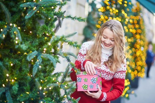 Cheerful young woman on a street of Paris decorated for Christmas, unpacking Christmas present