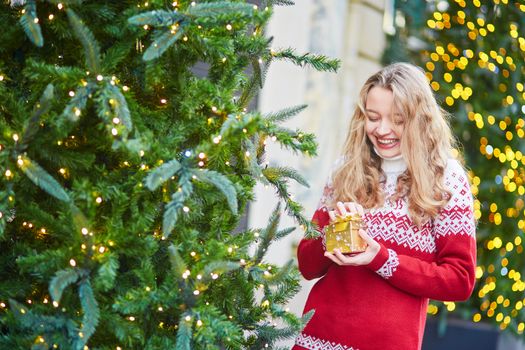 Cheerful young woman on a street of Paris decorated for Christmas, unpacking Christmas present