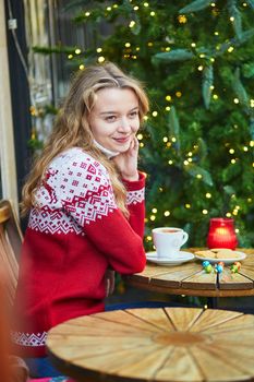 Cheerful young woman drinking hot chocolate in a cozy outdoor Parisian cafe decorated for Christmas