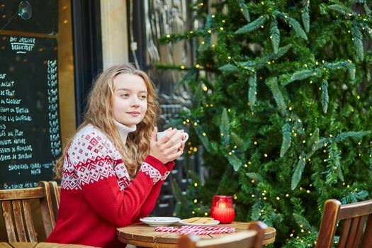Cheerful young woman drinking hot chocolate in a cozy outdoor Parisian cafe decorated for Christmas