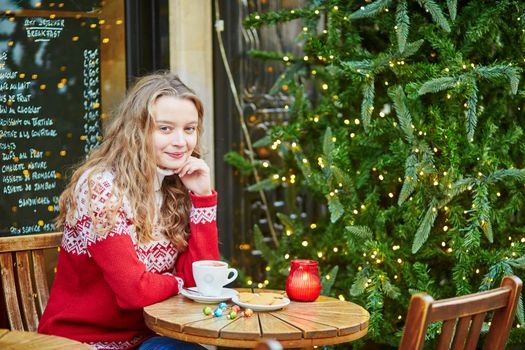 Cheerful young woman drinking hot chocolate in a cozy outdoor Parisian cafe decorated for Christmas