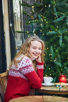 Cheerful young woman drinking hot chocolate in a cozy outdoor Parisian cafe decorated for Christmas