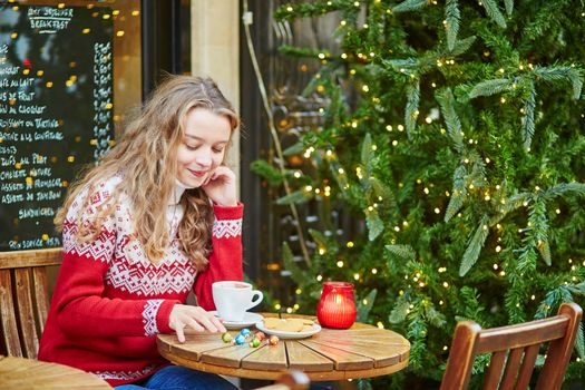 Cheerful young woman drinking hot chocolate in a cozy outdoor Parisian cafe decorated for Christmas