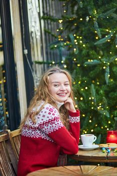 Cheerful young woman drinking hot chocolate in a cozy outdoor Parisian cafe decorated for Christmas