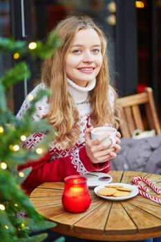 Cheerful young woman drinking hot chocolate in a cozy outdoor Parisian cafe decorated for Christmas