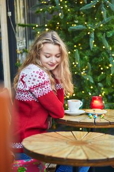 Cheerful young woman drinking hot chocolate in a cozy outdoor Parisian cafe decorated for Christmas