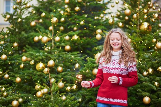 Cheerful young woman on a street of Paris decorated for Christmas with sparkling light in her hand
