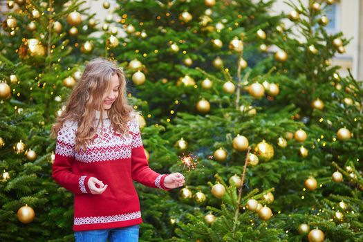 Cheerful young woman on a street of Paris decorated for Christmas with sparkling light in her hand