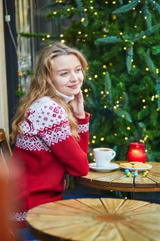 Cheerful young woman drinking hot chocolate in a cozy outdoor Parisian cafe decorated for Christmas