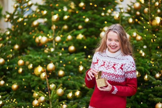 Cheerful young woman on a street of Paris decorated for Christmas, unpacking Christmas present