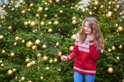 Cheerful young woman on a street of Paris decorated for Christmas with sparkling light in her hand