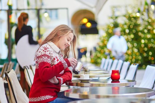 Cheerful young woman drinking hot chocolate in a cozy outdoor Parisian cafe decorated for Christmas