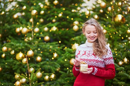 Cheerful young woman on a street of Paris decorated for Christmas, unpacking Christmas present