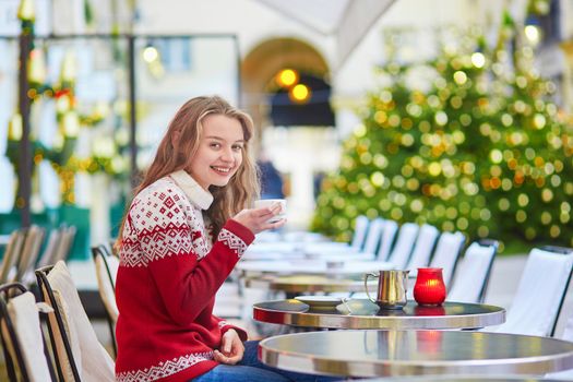 Cheerful young woman drinking hot chocolate in a cozy outdoor Parisian cafe decorated for Christmas