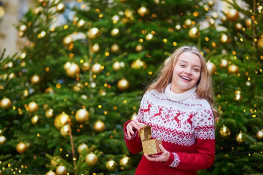 Cheerful young woman on a street of Paris decorated for Christmas, unpacking Christmas present