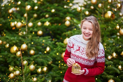 Cheerful young woman on a street of Paris decorated for Christmas, unpacking Christmas present