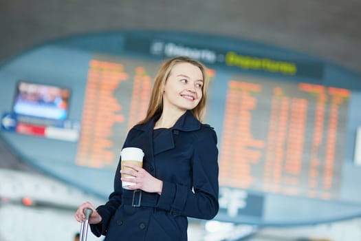 Young woman in international airport near the flight information board, with hot beverage to go