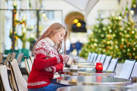Cheerful young woman drinking hot chocolate in a cozy outdoor Parisian cafe decorated for Christmas
