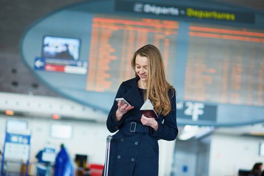 Young woman in international airport near the flight information board, holding French passport in her hand and using her mobile phone