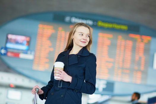 Young woman in international airport near the flight information board, with hot beverage to go