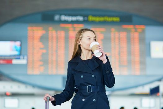 Young woman in international airport near the flight information board, with hot beverage to go