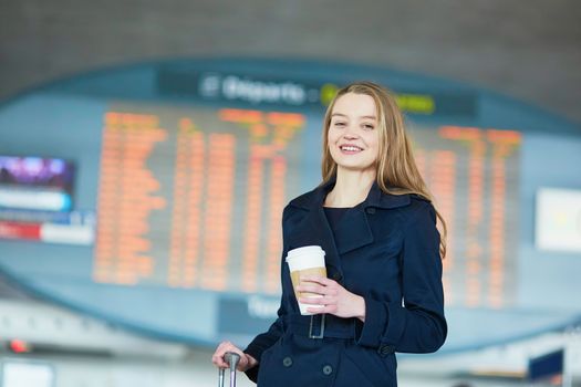 Young woman in international airport near the flight information board, with hot beverage to go