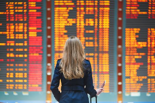 Young woman in international airport looking at the flight information board, holding passport in her hand, checking her flight